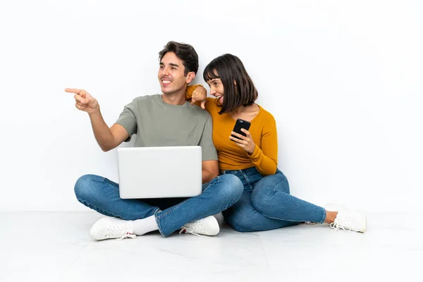 一对年轻的夫妇 带着笔记本电脑和手机坐在地板上 指着旁边 准备推出一款产品 — 图库照片