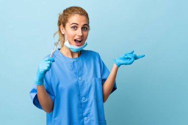 İzole edilmiş mavi arka planda aletleri tutan genç dişçi kadın şaşırdı ve parmağını yana çevirdi.