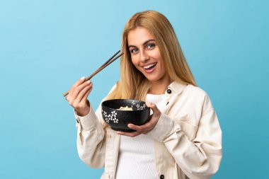 Genç Uruguaylı sarışın kadın izole edilmiş mavi arka planda elinde çubuklarla bir kase erişte tutuyor.