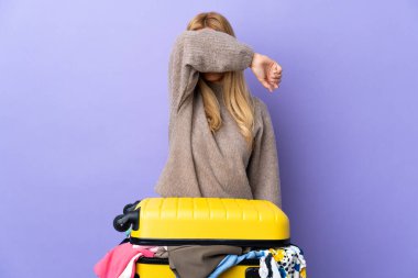 Genç Uruguaylı sarışın kadın bir bavul dolusu kıyafetle izole edilmiş mor duvarın üzerinden gözlerini elleriyle kapatıyor.