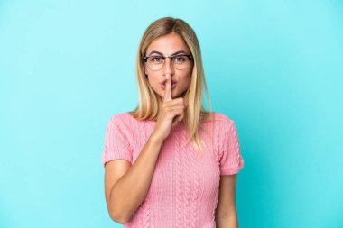 Sarışın Uruguaylı kız mavi arka planda izole edilmiş. Parmağını ağzına koyduğunda sessizlik işareti gösteriyor.