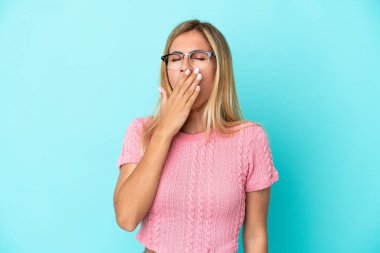 Sarışın Uruguaylı kız mavi arka planda izole edilmiş esniyor ve ağzını eliyle açık tutuyor.