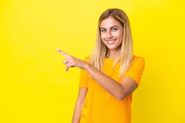 Sarışın Uruguaylı kız, sarı arka planda izole edilmiş bir şekilde bir ürün sunmak için kenarı işaret ediyor.