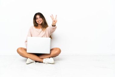 Yerde dizüstü bilgisayarı olan genç bir kadın beyaz arka planda izole edilmiş mutlu ve parmaklarıyla üçe kadar sayıyor.