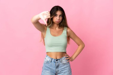 Pembe arka planda izole edilmiş genç beyaz kadın olumsuz ifadeyle baş parmağını aşağı gösteriyor.