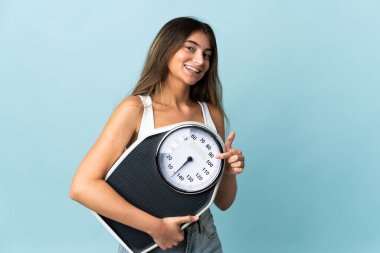 Mavi arka planda izole edilmiş beyaz bir kadın elinde bir tartı makinesi tutuyor ve ona doğrultuyor.