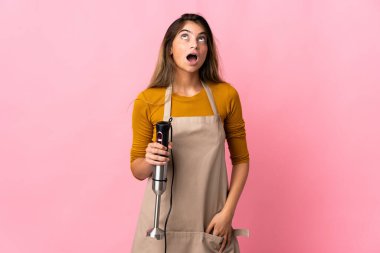 Genç aşçı kadın el blenderi kullanıyor pembe arka planda izole edilmiş yukarı bakıyor ve şaşırmış bir ifadeyle