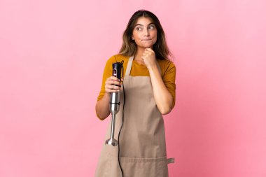 Genç aşçı kadın pembe arka planda izole edilmiş el blenderi kullanıyor. Şüpheleri ve düşünceleri var.