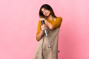 Genç aşçı kadın el blenderi kullanıyor. Pembe arka planda izole edilmiş. Çabaladığı için omuz ağrısı çekiyor.