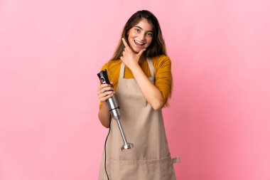 Genç aşçı kadın pembe arka planda izole edilmiş el blenderi kullanıyor mutlu ve gülümsüyor.