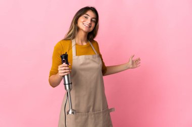 Genç aşçı kadın el blenderi kullanıyor. Pembe arka planda izole edilmiş. Gelmesi için ellerini yana uzatıyor.