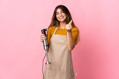 Pembe arka planda el blenderi kullanan genç aşçı kadın para hareketi yapıyor.