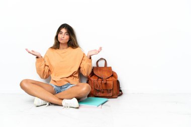 Yerde dizüstü bilgisayarlı genç bir bayan oturuyor. Beyaz arka planda izole edilmiş. El kaldırırken şüpheleri var.