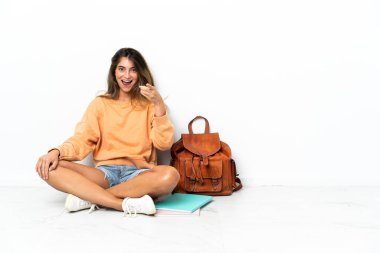 Yerde dizüstü bilgisayarlı genç bir bayan oturuyor. Beyaz arka planda izole edilmiş. Şaşırmış ve ön tarafı işaret ediyor.