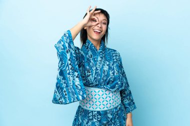 İzole edilmiş mavi arka planda kimono giyen genç bir kadın parmaklarıyla tamam işareti gösteriyor.