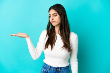 Mavi arka planda izole edilmiş genç beyaz bir kadın. Kopyalama alanını şüphe ile tutuyor.