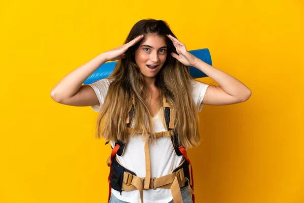 驚きの表情で黄色の背景に隔離された大きなバックパックを持つ若い登山家の女性 — ストック写真