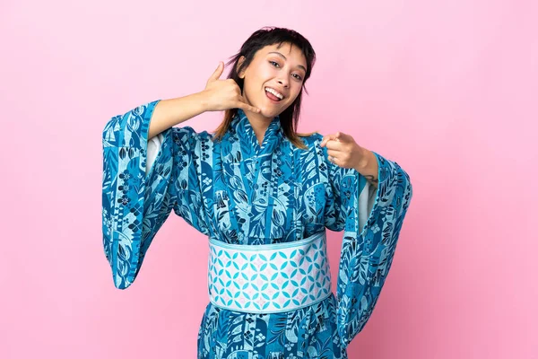 İzole edilmiş mavi arka planda kimono giyen genç bir kadın telefon hareketinde bulunuyor ve ön tarafı işaret ediyor.