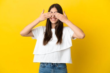 Sarı arka planda izole edilmiş genç beyaz bir kadın. Gözleri elleriyle kaplıyor ve gülümsüyor.