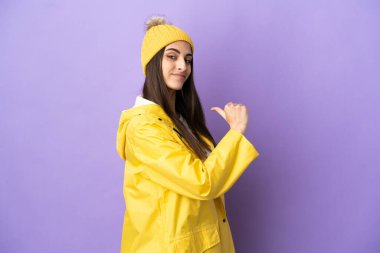 Mor arka planda izole edilmiş, yağmura dayanıklı bir ceket giyen beyaz bir kadın gururlu ve kendini beğenmiş.
