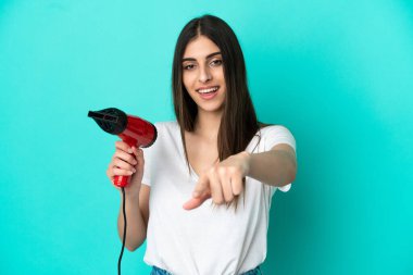 Mavi arka planda saç kurutma makinesini tutan beyaz bir kadın kendine güvenen bir ifadeyle seni işaret ediyor.