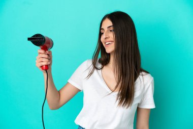 Kafkasyalı genç bir kadın elinde saç kurutma makinesiyle mavi arka planda mutlu bir ifadeyle izole edilmiş.