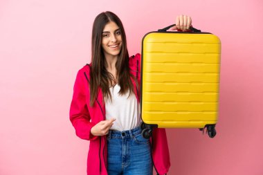 Pembe arka planda izole edilmiş genç beyaz kadın seyahat valiziyle geziyor.