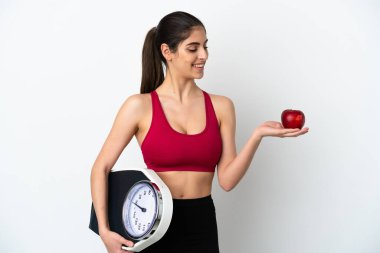 Beyaz arka planda izole edilmiş genç beyaz bir kadın elmaya bakarken bir tartı makinesi tutuyor.