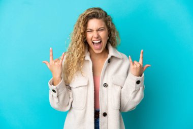 Mavi arka planda izole edilmiş genç sarışın kadın boynuz hareketi yapıyor.