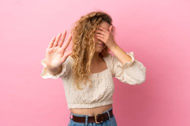 Pembe arka planda izole edilmiş genç sarışın kadın dur hareketi yapıyor ve yüzünü kapatıyor.