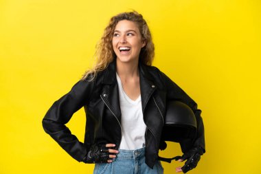 Sarı arka planda izole edilmiş motosiklet kasklı genç sarışın kadın kolları kalçasında ve gülümsüyor.