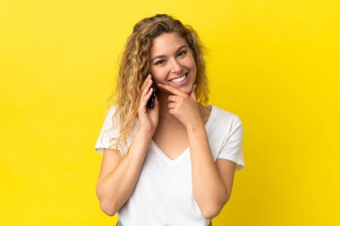 Sarı arka planda izole edilmiş cep telefonu kullanan genç sarışın kadın mutlu ve gülümsüyor.