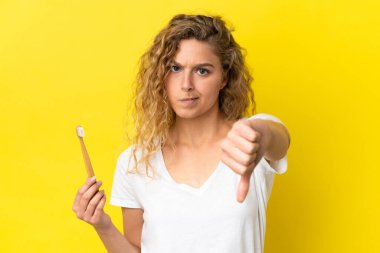 Sarı arka planda dişlerini fırçalayan genç beyaz bir kadın olumsuz bir ifadeyle baş parmağını gösteriyor.