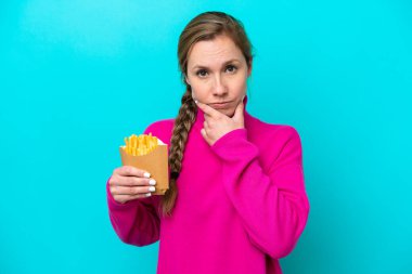 Elinde kızarmış cips tutan beyaz bir kadın mavi arka planda izole edilmiş olarak düşünüyor.