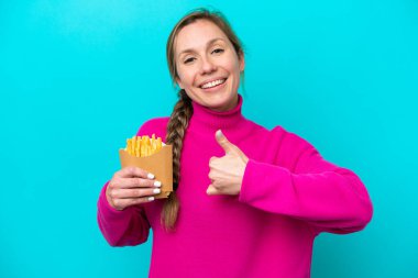 Elinde kızarmış cips tutan beyaz bir kadın mavi arka planda izole edilmiş bir şekilde baş parmak işareti yapıyor.