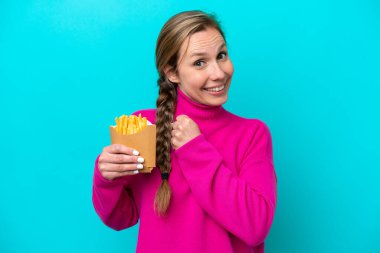 Mavi arka planda izole edilmiş kızarmış patates kızartması tutan beyaz bir kadın zaferi kutluyor.