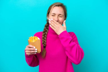 Mavi arka planda izole edilmiş kızarmış patates kızartması tutan genç beyaz kadın mutlu ve ağzı kapalı gülümsüyor.