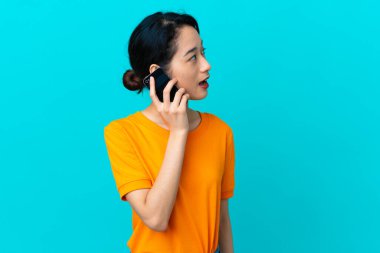 Genç Vietnamlı kadın mavi arka planda izole edilmiş cep telefonuyla birileriyle sohbet ediyor.