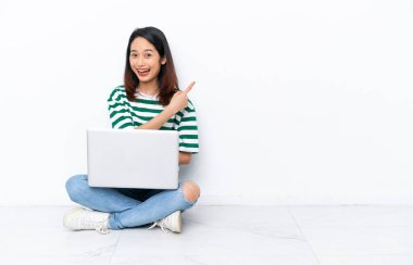 Yerde dizüstü bilgisayarı olan genç Vietnamlı kadın beyaz duvarda izole edilmiş, şaşırmış ve işaret ediyor.