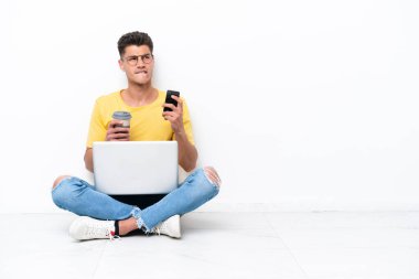 Genç adam beyaz arka planda izole bir şekilde yerde oturuyor. Elinde kahve ve bir şeyler düşünürken cep telefonu var.