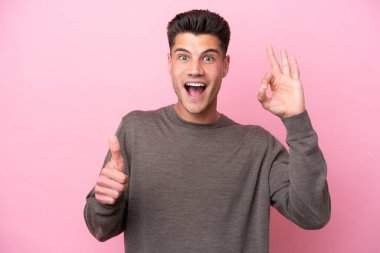 Pembe arka planda izole edilmiş genç beyaz bir adam tamam işareti ve baş parmak hareketi gösteriyor.