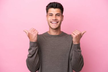 Pembe arka planda izole edilmiş genç beyaz bir adam. Baş parmakları havada ve gülümsüyor.