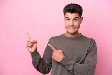 Pembe arka planda izole edilmiş genç beyaz adam korkmuş ve kenarı işaret ediyor.