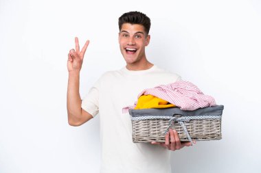 Beyaz arka planda izole edilmiş çamaşır sepetini tutan genç beyaz adam gülümsüyor ve zafer işareti gösteriyor.