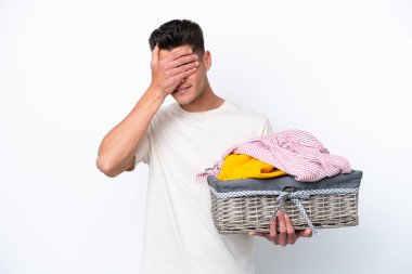 Çamaşır sepetini tutan Kafkasyalı genç adam beyaz arka planda izole edilmiş, yorgun ve hasta bir ifadeyle.