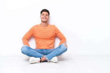 Genç adam beyaz arka planda izole bir şekilde yerde oturuyor. Kolları kalçasında ve gülümsüyor.