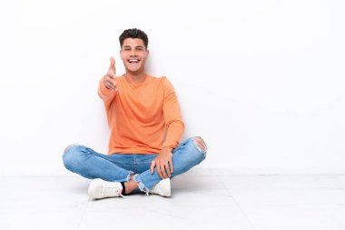 Genç adam beyaz arka planda izole bir şekilde yerde oturuyor ve iyi bir anlaşma için el sıkışıyor.