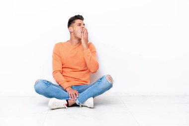Genç adam beyaz arka planda izole bir şekilde yerde oturuyor esniyor ve ağzını eliyle açık tutuyor.