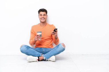 Genç adam beyaz arka planda izole bir şekilde yerde oturuyor. Elinde kahve ve cep telefonu var.