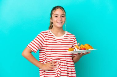 Mavi arka planda, kolları kalçasında poz veren ve gülümseyen waffle tutan beyaz bir kız.